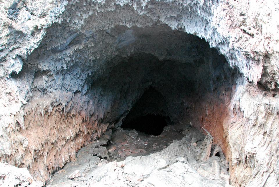 Photo: An entrance to one of the lava tubes. The temperature of the air here was measured at 170 °C. The blue coating on the lava stalactites are salts that have precipitated as the hot air mixes with the outside atmosphere.