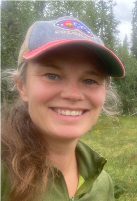 Picture of the PhD candidate smiling. She is wearing a red and blue cap a green jacket.