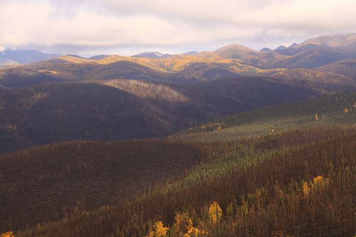 A hilly landscape photo with a partly cloudy sky. The photo appears to be taken from the top of a tall hill and features lower hills to horizon, covered by boreal forest. 