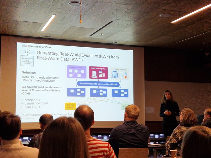 A lecture audience watches a presentation on generating real‑world evidence from real‑world data. The projected slide contains diagrams showing data sources, transformation into a common model, and standardized analytics. A presenter stands beside the screen.