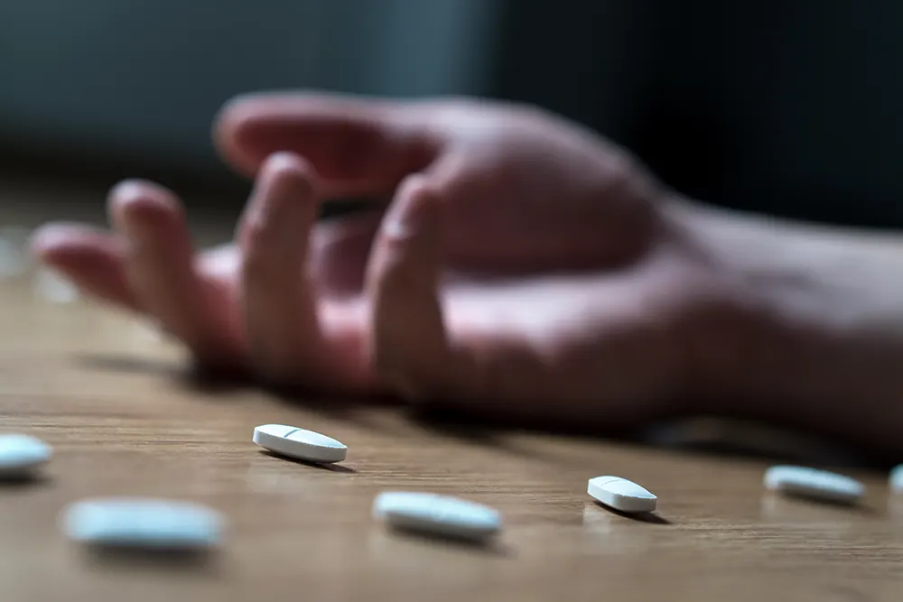Tablettar som ligg på eit golv. I bakgrunnen ligg ei slapp hand. Resten av kroppen utanfor biletet