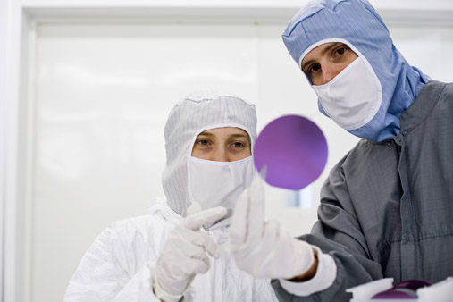 Two researchers dressed in full protective lab clothes including face mask. One is holding up a circular purple solar wafer that they both are examining. 
