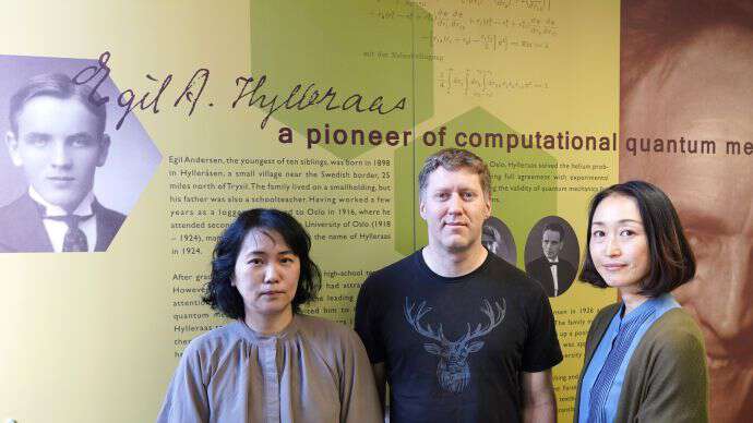 Three persons standing in front of a board presenting the researcher Hylleraas. The persons are who ladies and one man.
