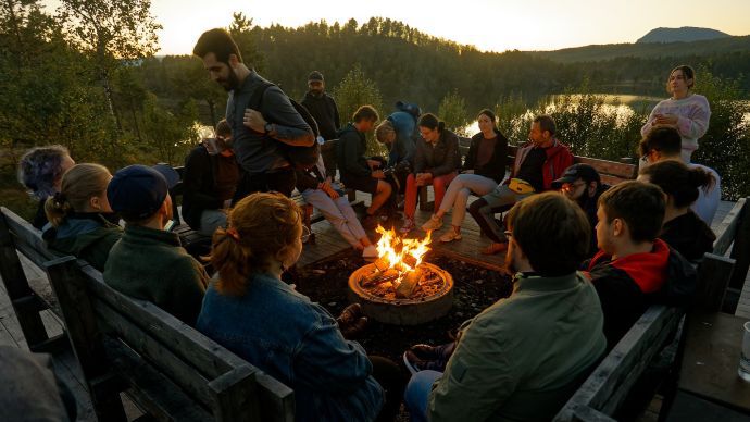 Photo showing young adults around a bonfire i nature by the lake. Hylleraas members enjoying a barbecue by a lake at Malangen resort..
