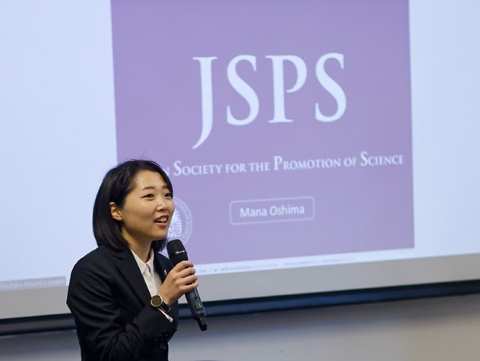 Young woman from Japan is having av short speech in front of a slide with the writing "JSPS". 
