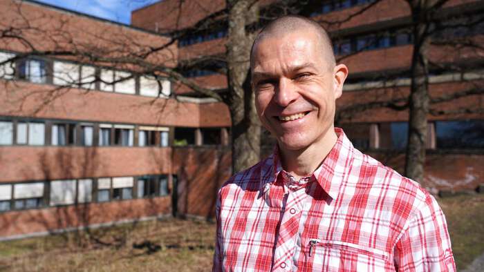 Smiling and a happy man with a red and white shirt standing outside av red-ish building. 