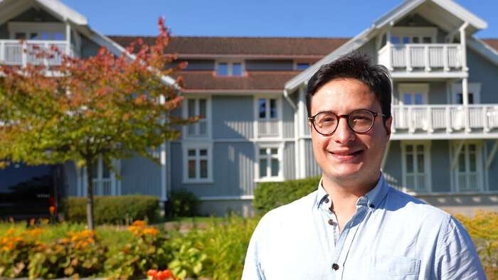 Young man whith a blue shirt and glasses, standing outside Losby gods, a historic manor hotel. surrounded by threes in summertime. 