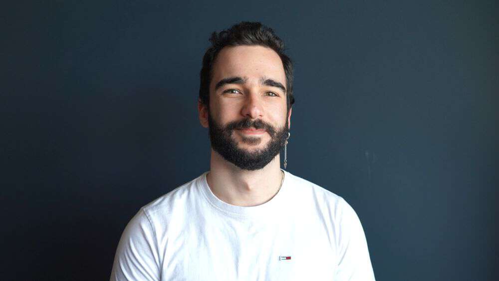 A young man, smiling, with a white t-short in front of a blue wall. 