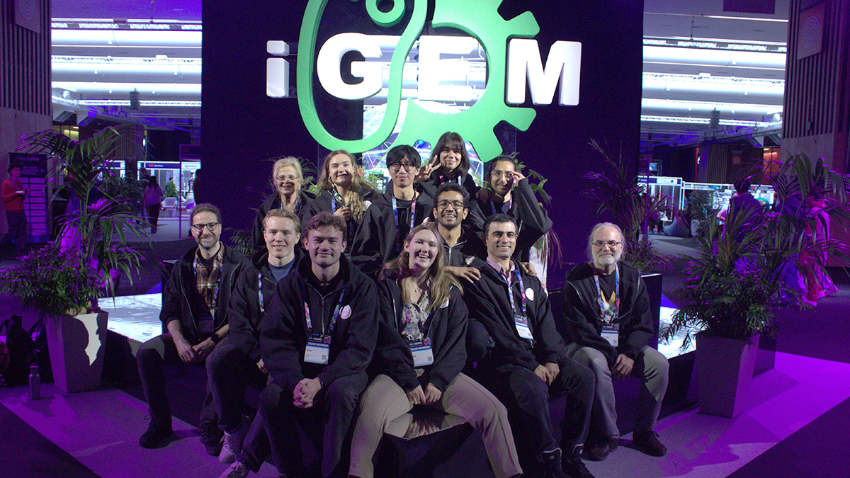 Group photo of the 2025 UiO iGEM-team; sitting in from of a large, glowing iGEM-logo.