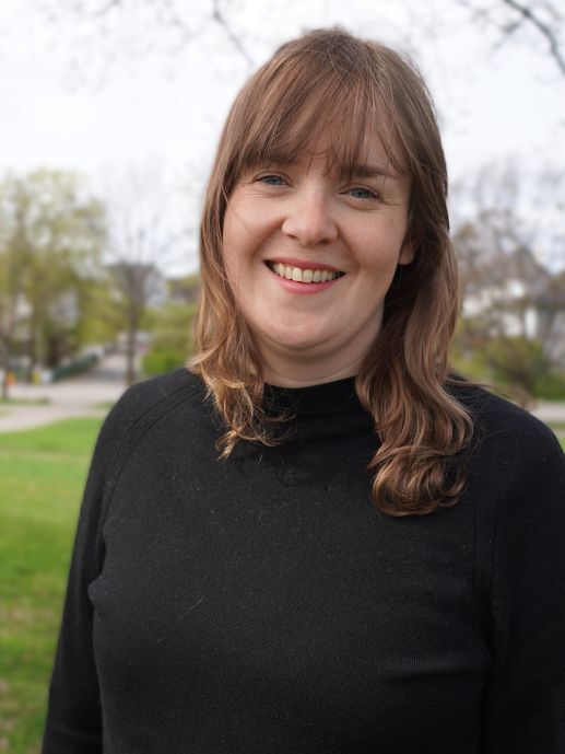Young woman with black jumper, brown hair, blue eyes. Outside on the green. 