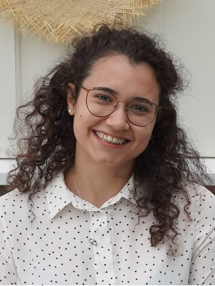 Image may contain: A person (Marinella) is smiling and looking straight at the camera. They have long brown hair and are wearing a white shirt with shirt with black dots.