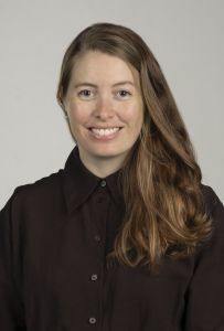 Image may contain: A person (Julie Hessevik) looking straight at the camera. The person has long brown hair covering the left shoulder and is wearing a black collared shirt.