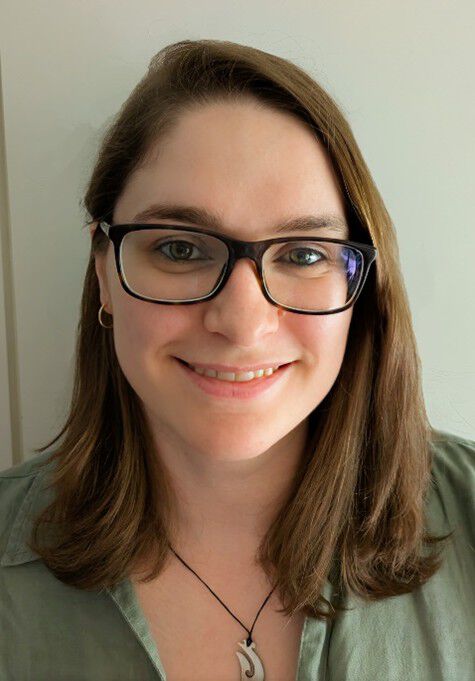 Image may contain: A person (Sara) looking straight at the camera. They have shoulder-length brown hair and are wearing a green shirt and glasses.