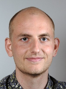 Image may contain: A person (Thomas) looking straight at the camera. They have a shaved head and are wearing a spotted blue, white and yellow shirt.