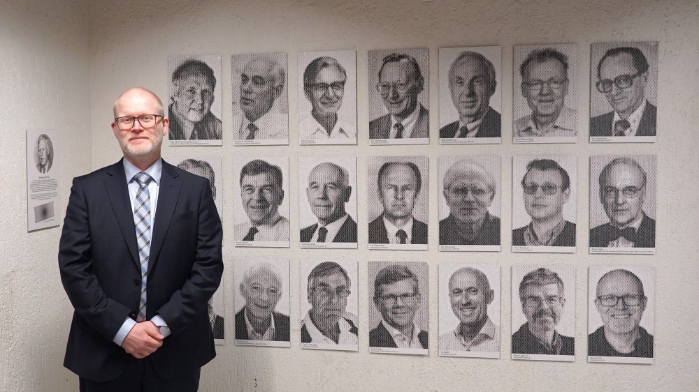 Portretter i sort-hvitt, 21 stk. Til venstre står en mann i mørk dress og smiler. 