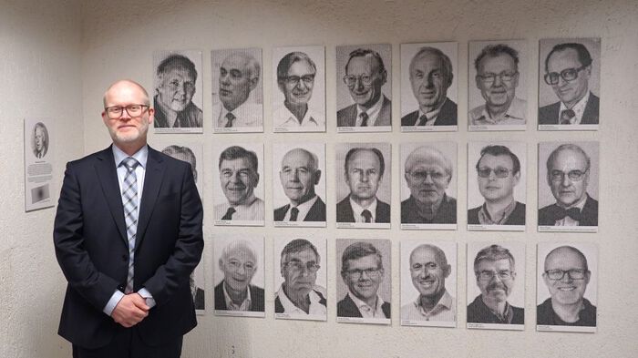 Portretter i sort-hvitt, 21 stk. Til venstre står en mann i mørk dress og smiler. 