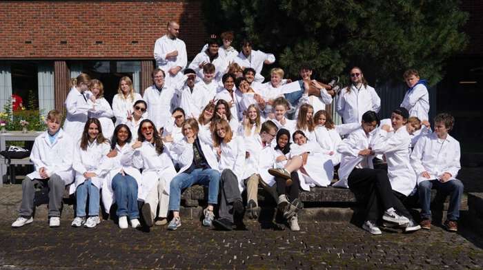 Unge studenter med labfrakk sitter lett kaotisk på benker foran en murbygning. 