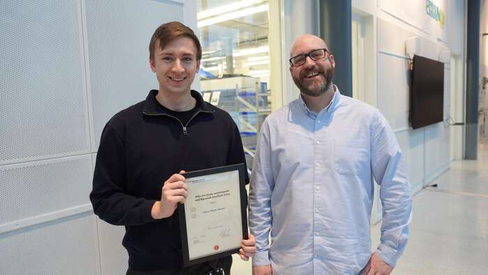 Oliver Moen-Duran fikk prisen for beste masteroppgave sammen og et gavekort fra Kjemisk institutt. Her sammen hovedveilederen, Simon Philip Cooil. På bildet står de i en lys korridor hvor det er mulig å se gjennom et vindu med kjerneinfrastruktur. 
