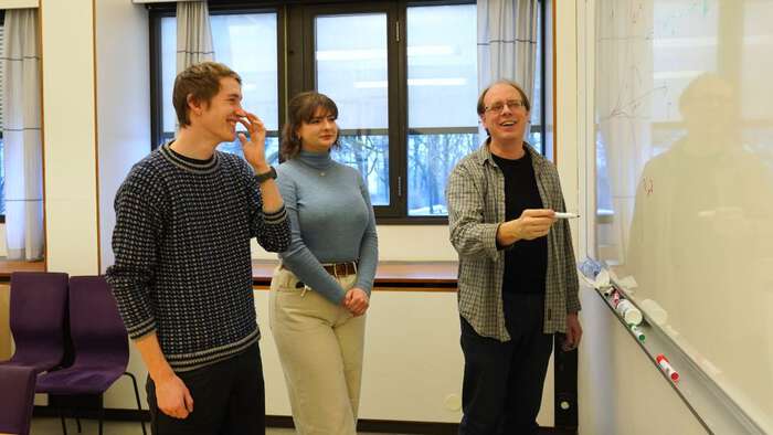 Bildet viser to studenter, en ung mann og en ung kvinne i dialog med en mannlig underviser. 