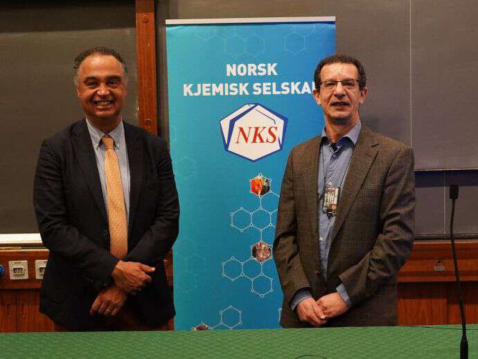 To menn smiler med en skilt bak hvor det står Norsk Kjemisk Selskap. Mennene er professor Jérôme Lacour og 2) Professor Mohamed Amedjkouh.