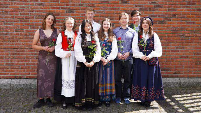 8 studenter, 3 gutter og 5 jenter står med rose og i finklær eller bunad foran en rød murvegg.