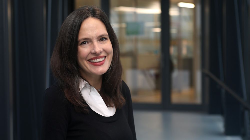 Smiling woman with dark hair looking at the camera. In the background, there is a glass door to a building.