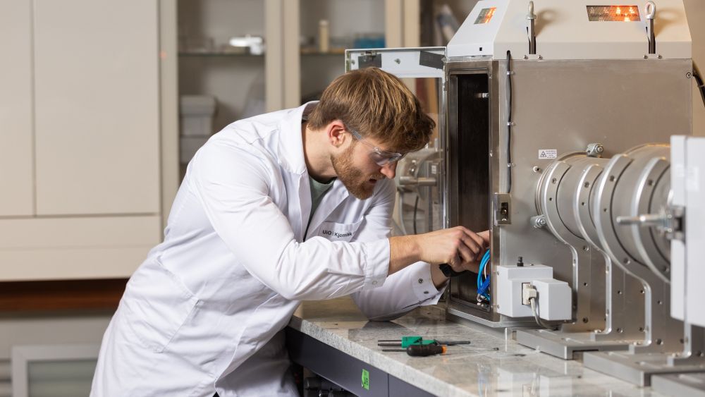 Ung mannlig forsker som gjør justeringer på et stort og avlangt instrument innen nanoteknologi. Han har på seg labfrakk og vernebriller, og er på et laboratorium.