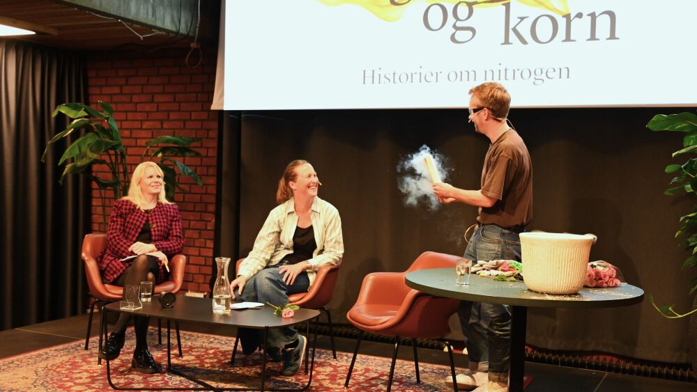 To kvinner sitter på scene, mann holder rykende bok