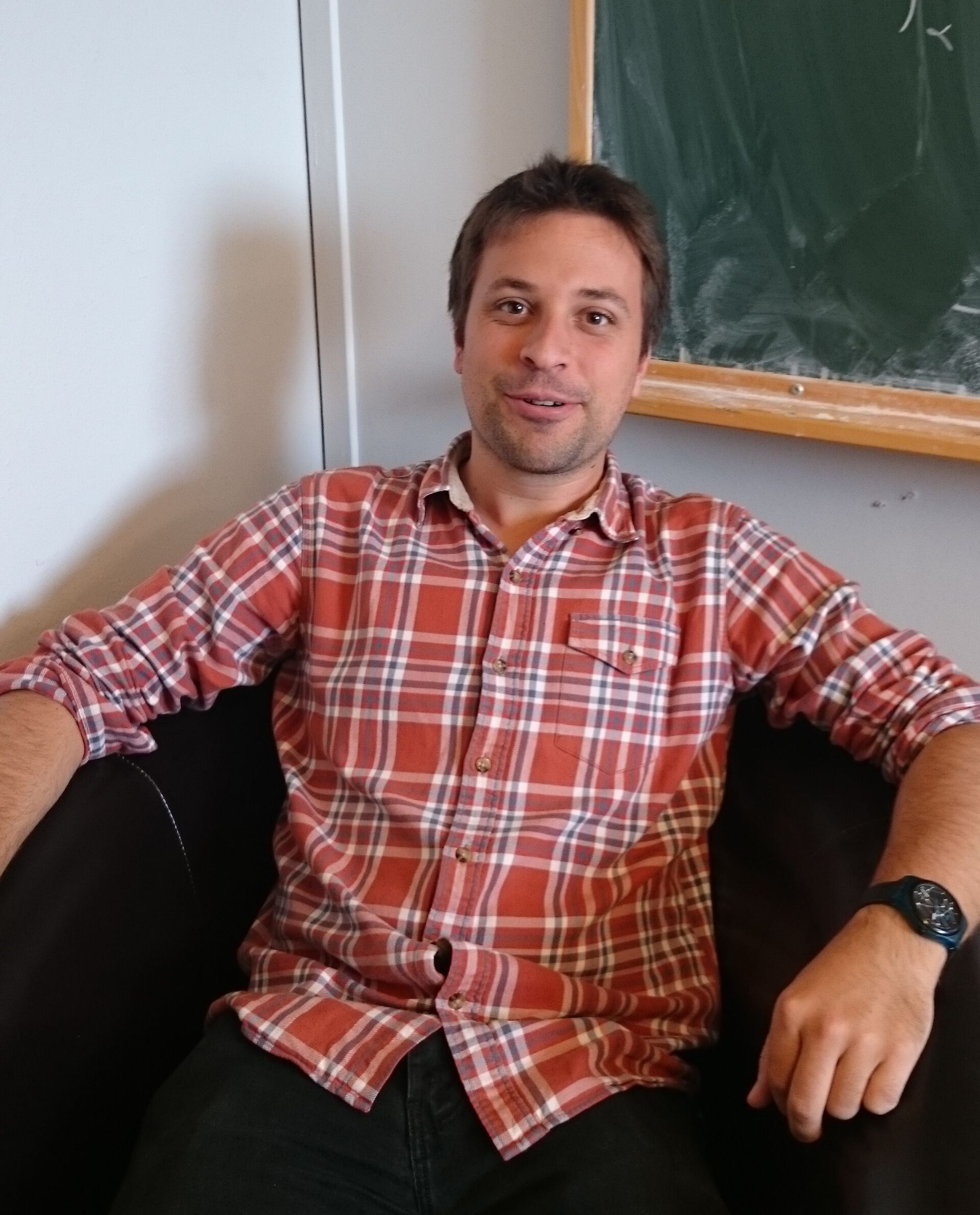 Fabian Barras, a man with brown hair and short beard, wearing a plaid shirt and sitting in a chair. 