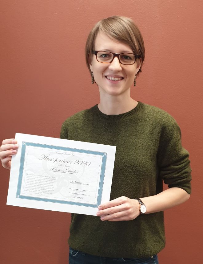 A woman with light hair, wearing a green sweater and glasses and holding an award. 