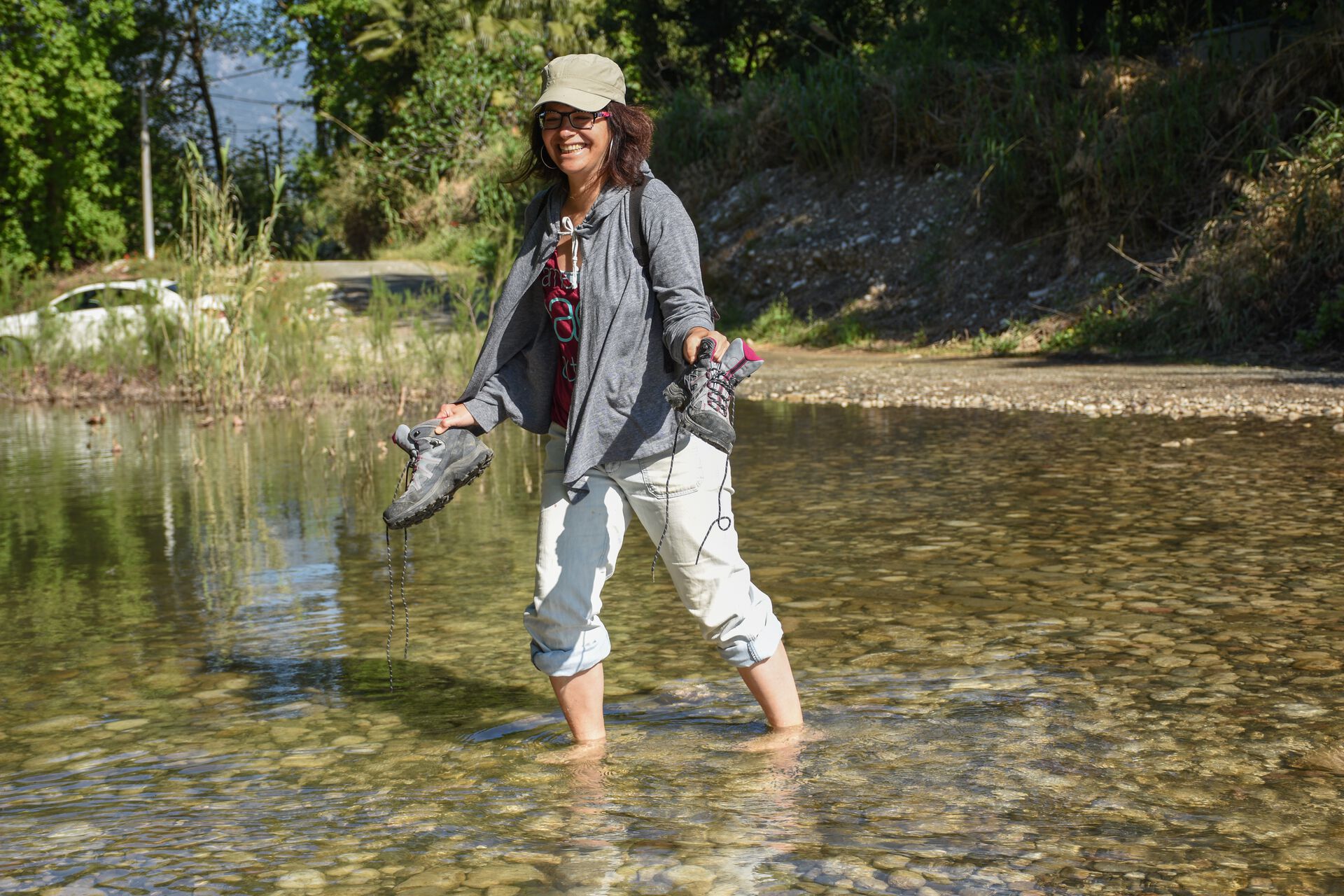 A woman in  field clothes, crossing a river barefoot and carrying her shoes.  