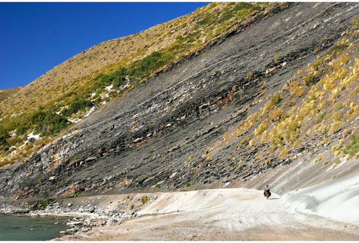 A hillside covered with rocks and yellow grass takes up most of the photo, and at the bottom there is a dirt road where a loan biker can be seen from afar.