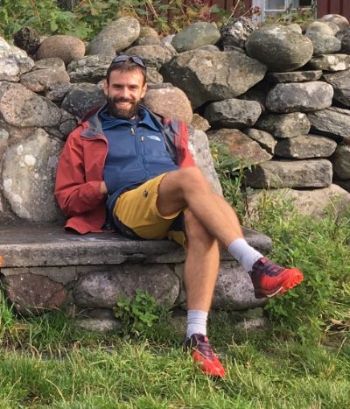A man wearing outdoor clothes and sitting on a stone bench in front of a stone wall. 