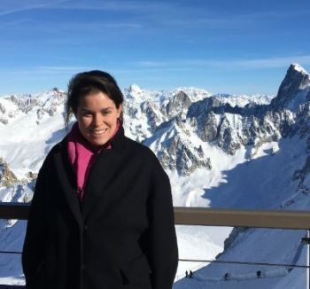 A woman in pink sweater and black jacked standing in front of a wooden fence, and a mountain range covered with snow and clear sky in background.