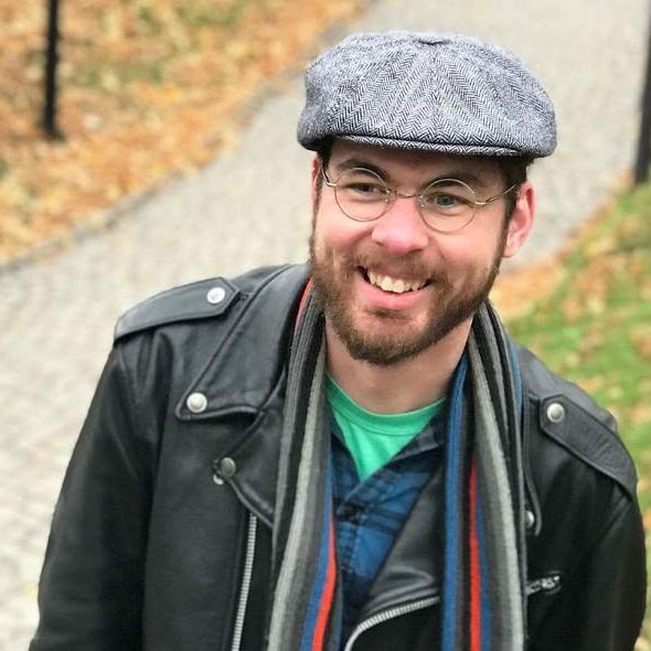 A photo of a man with brown hair and beard, smiling and wearing a leather jacket, plain cap, glasses and striped scarf. 
