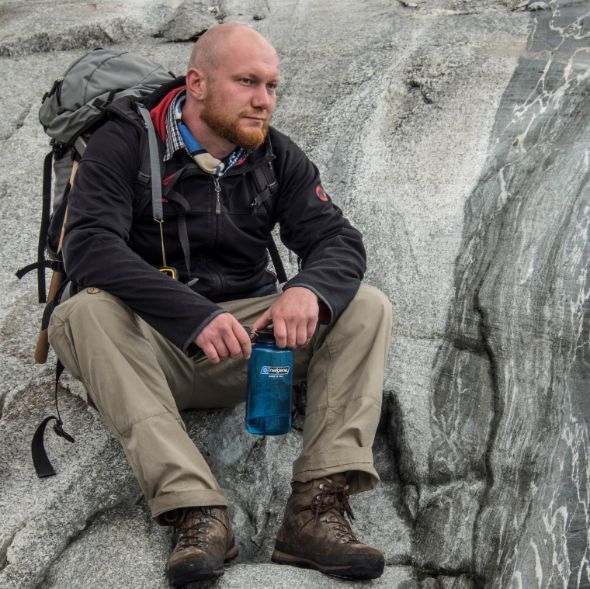 Man sitting on grey rock. The man is bald and with a short, red beard. He is wearing a black jacket with a short underneath and a grey backpack. He has beige pants and is holding a blue bottle. On his feet he has brown hiking boots. His head is looking slightly of the right of the camera.