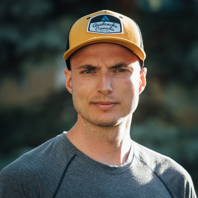 Close-up of smiling man. He is wearing a yellow cap with a black and white logo and a grey sweater. The background is blurry.