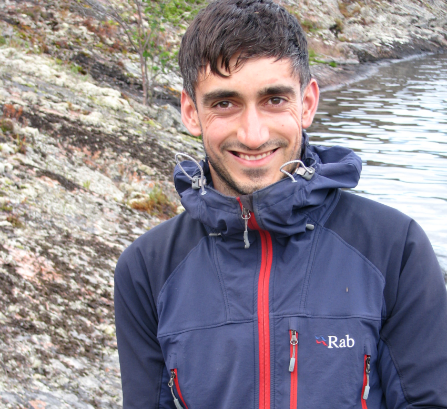 A man in outwear standing in front of a lake and smiling. 