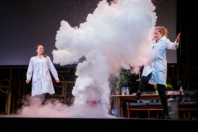 Bildet inneholder to jenter i labfrakk og vernebriller i en røyksky.