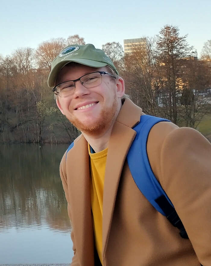 A picture of Björn Heyn. He has a green cap on, thin-rimmed glasses, a red beard and a brown coat.