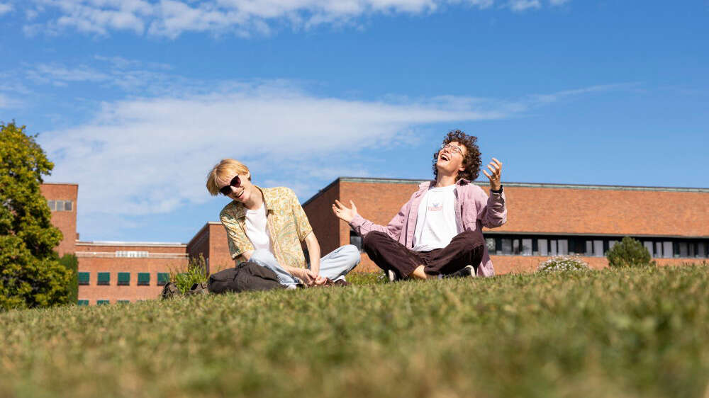 To studenter sitter på gresset med beina i kors og ser glade ut; universitetsbygg i mur i bakgrunn